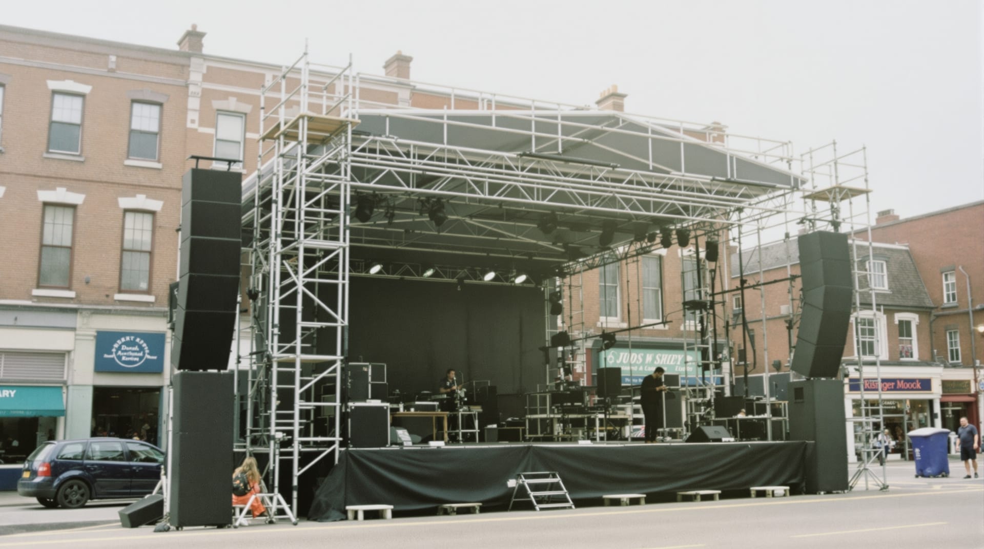 A small outdoor stage set up in a closed city street with speaker stacks and a lighting truss